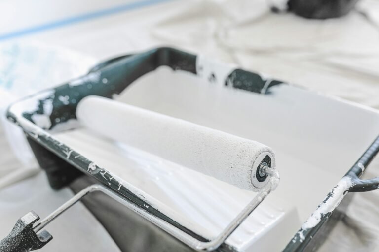 A white paint roller rests in a dark tray of white paint on a protective sheet.