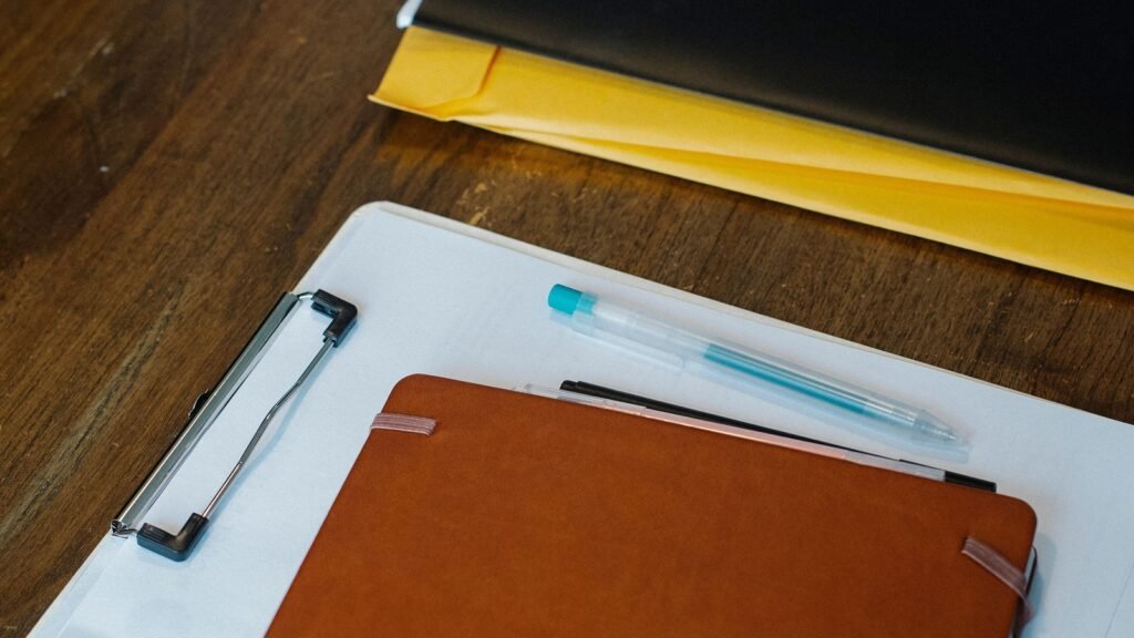 A notebook and pens rest on a blank clipboard beside stacked folders on a wooden desk.