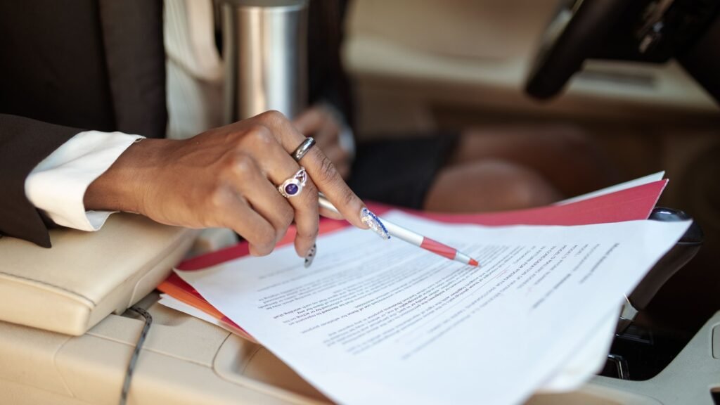 A hand holding a pen over papers and a red folder inside a car.