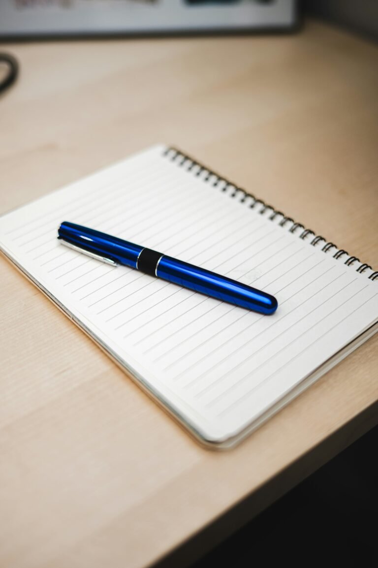 Blue pen resting diagonally on a blank lined notebook on a wooden desk.