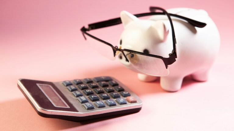 A white piggy bank wearing glasses beside a calculator on a pink surface.