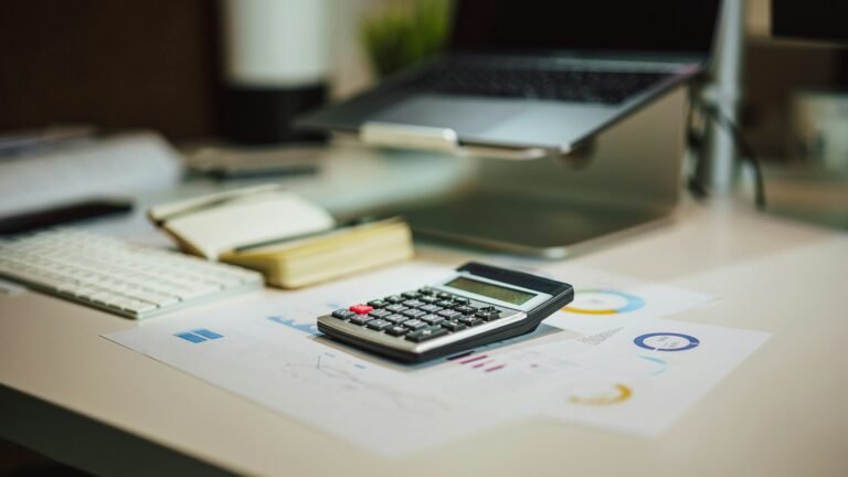 Calculator on printed charts beside a laptop and notebook on a desk.