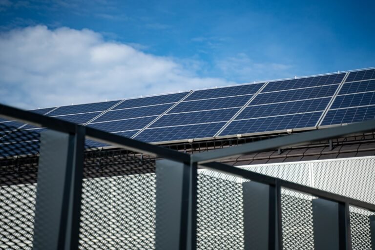 Blue solar panels on a roof beneath a partly cloudy sky, with a metal railing in the foreground.