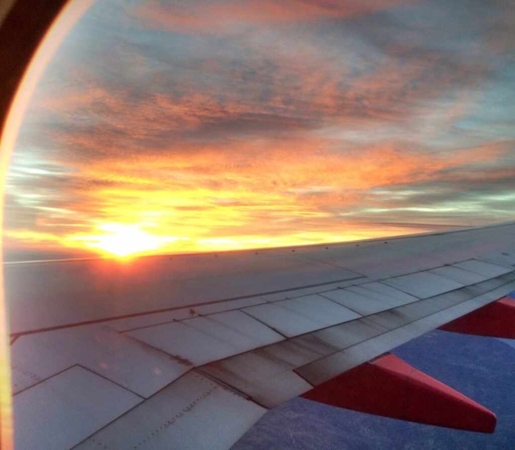An airplane wing beneath orange sunset clouds, viewed through a window.