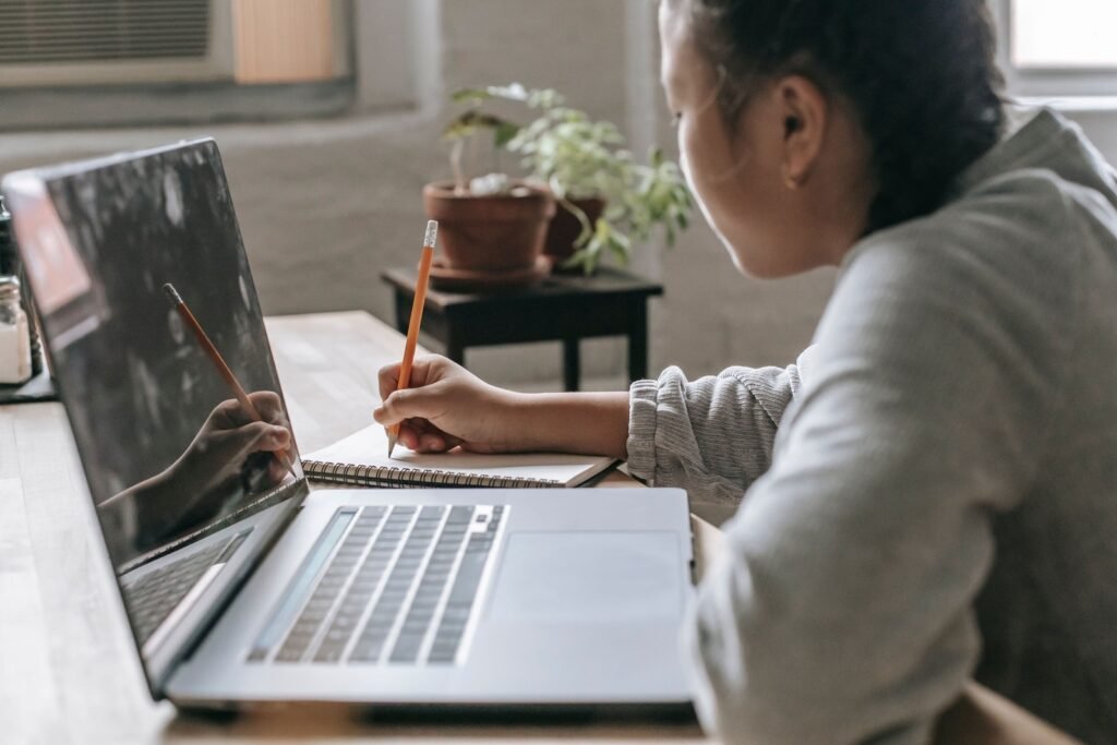 A person writes with a pencil in a spiral notebook beside an open laptop on a wooden desk.