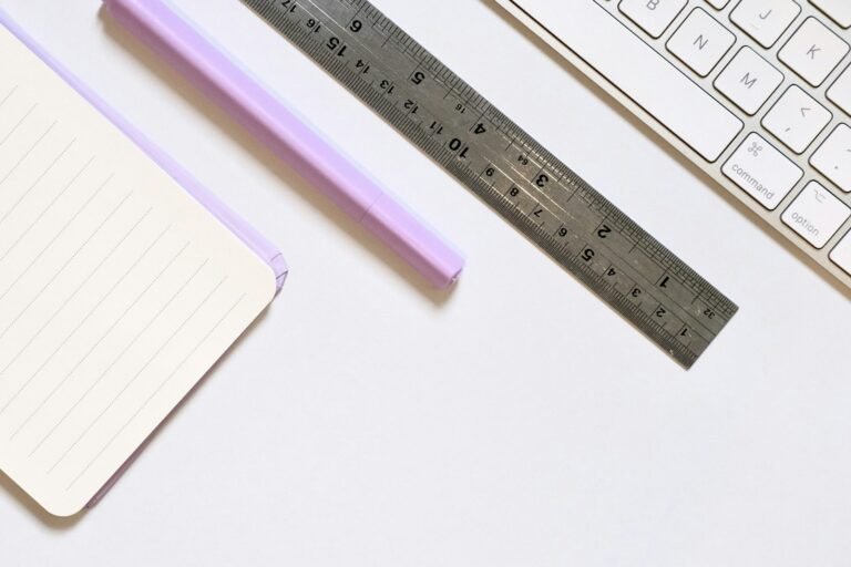 A blank notebook, purple pen, metal ruler and keyboard on a white desk.