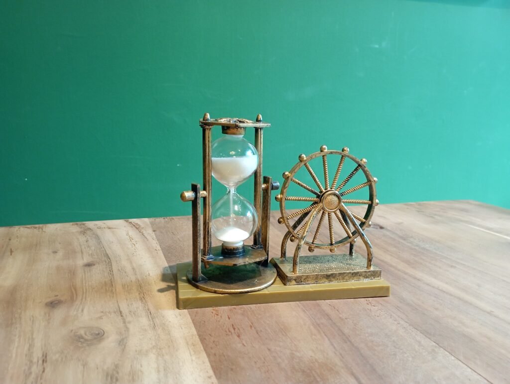 A glass hourglass and metal wheel ornament on a wooden table.