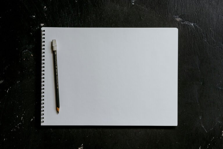 A pencil rests on a blank spiral notebook on a dark textured table.