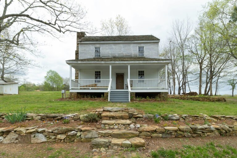 A white two-story country house with a front porch, stone steps and surrounding trees.