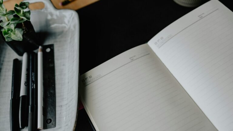 An open blank notebook beside pens, a pencil and a ruler.