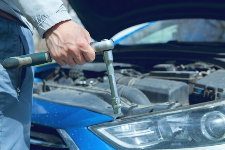 A hand holds a socket wrench above an open car engine bay.