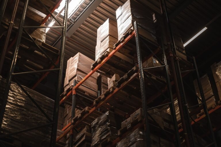 Low-angle view of stacked cartons on tall warehouse racks beneath roof lights.