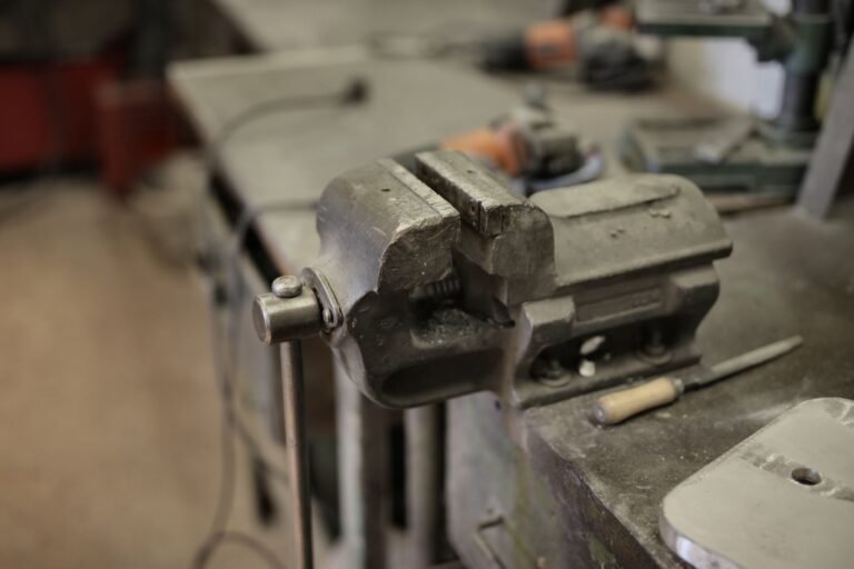 A metal bench vise beside a hand file, with power tools blurred in the background.